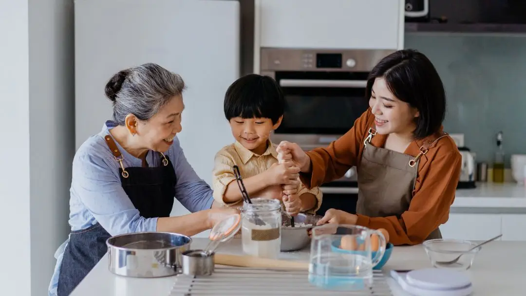 Grandparent, child, and adult cooking together in a modern kitchen, representing family bonding and the importance of protecting home and lifestyle in Singapore.